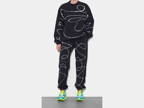 Model is shown wearing 100% cotton black sweatsuit, perfectly cut, which has scrawled, drippy white hand-painted line running across the body, sleeves, and down each leg.